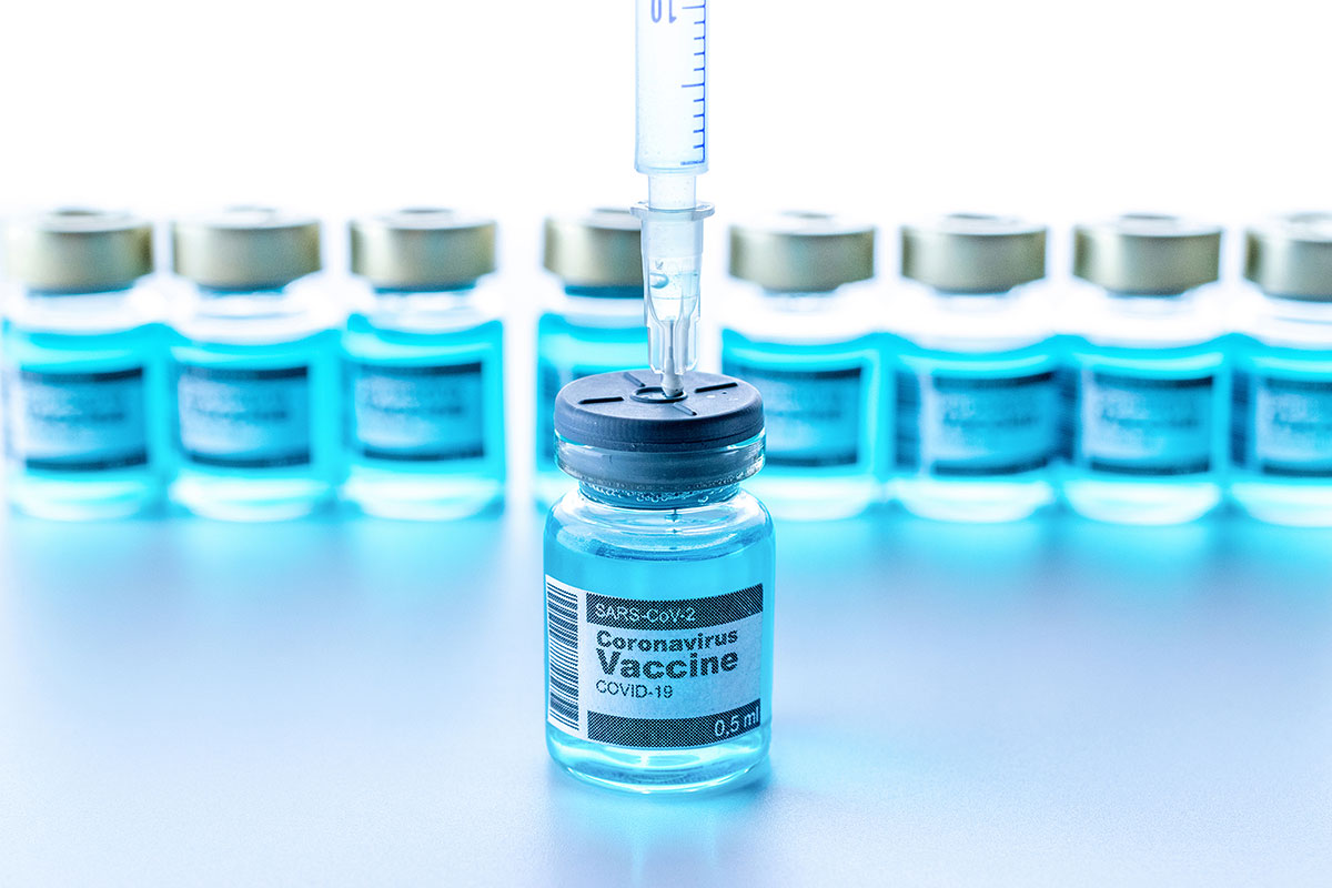 Syringe is filled with Covid-19 vaccine liquid from vial. Eight more vials positioned blurred in the background