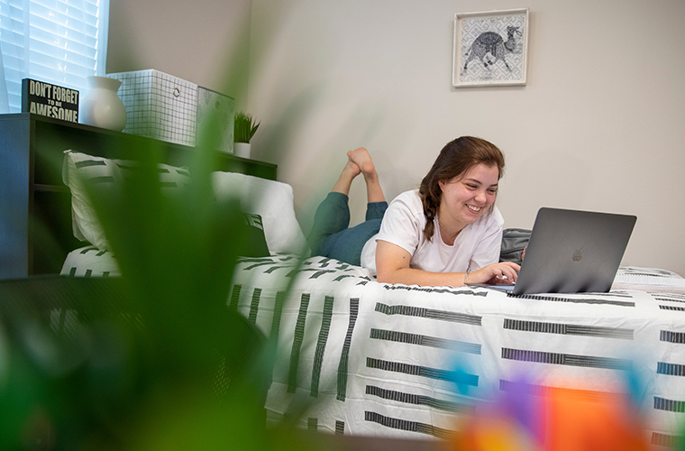 Female student lays in dorm bed, smiling while looking at laptop