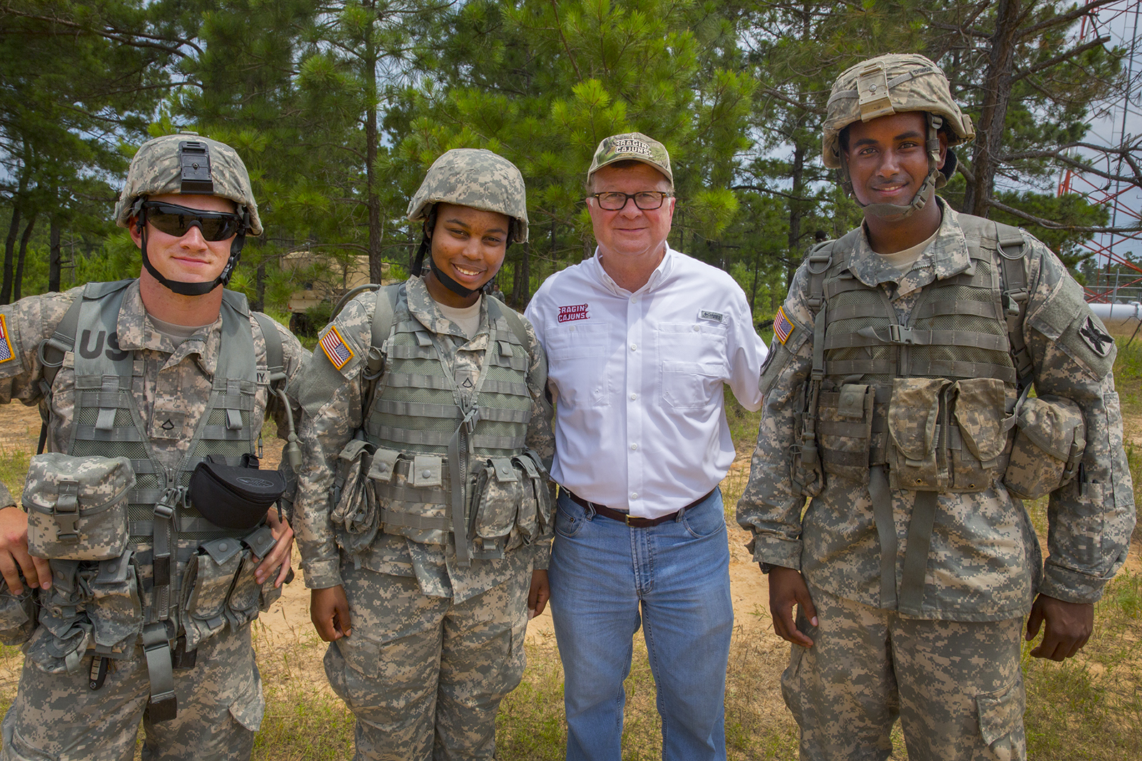 3 military students in uniform stand smiling next to a man in a white button-down shirts and blue jeans with a camouflage hat with white lettering "Ragin Cajuns"