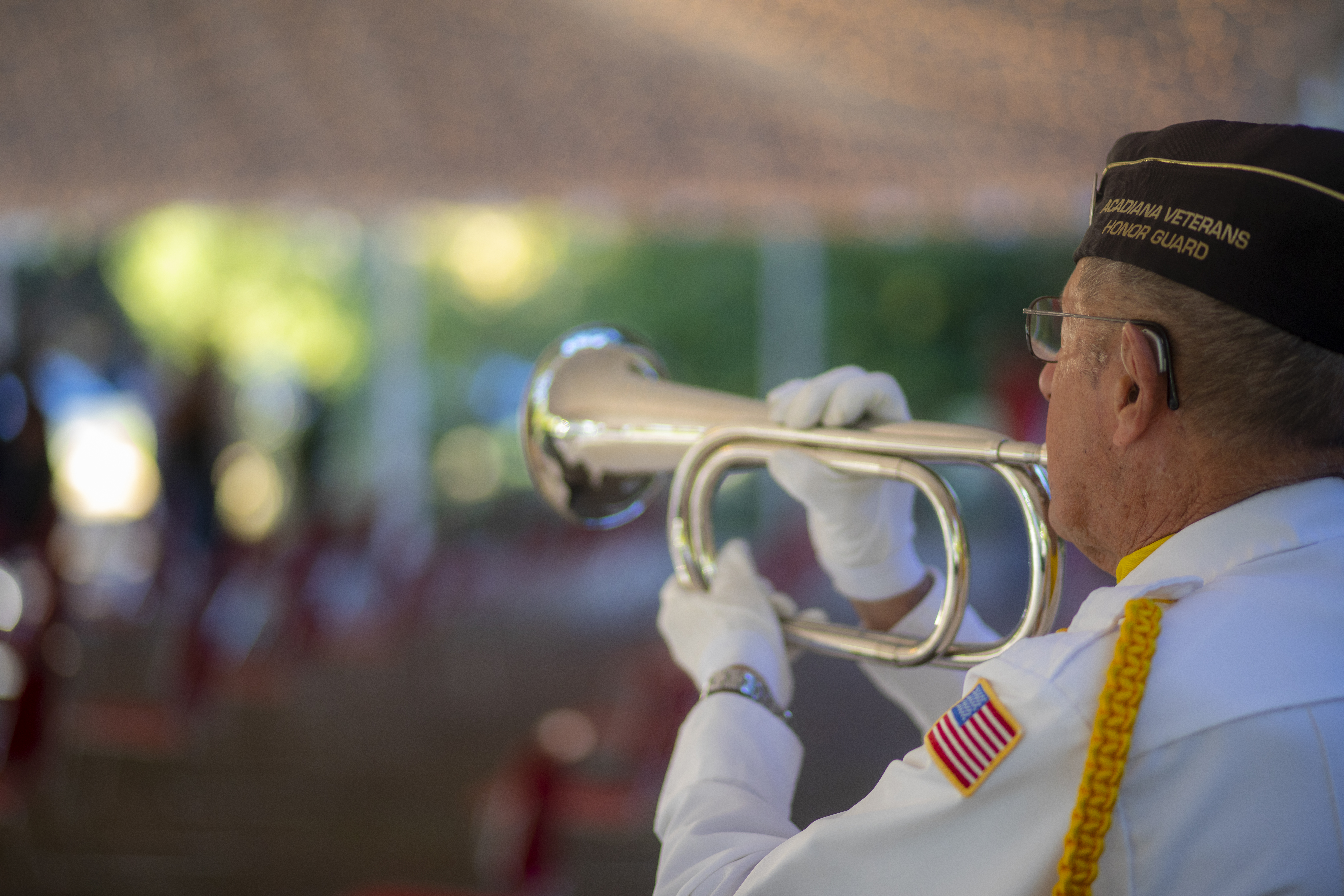 Elderly veteran pictured from behind playing the trumpet, wearing brown cap with gold text "Acadiana Veterans Honor Guard"