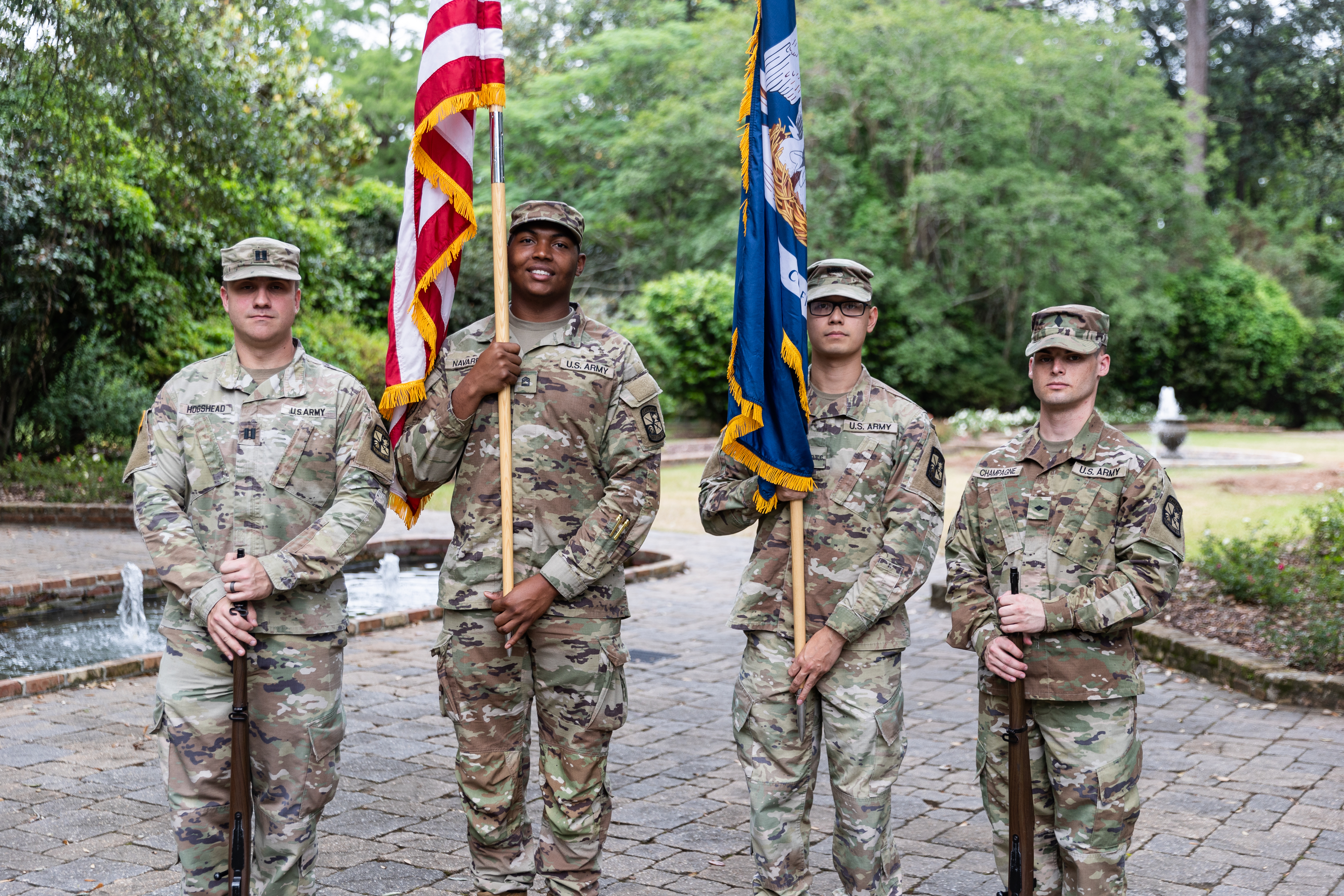 Four U.S. Army students stand. The two outermost hold rifles, while two in the center hold flags - to the left an American flag, to the left the Louisiana state flag