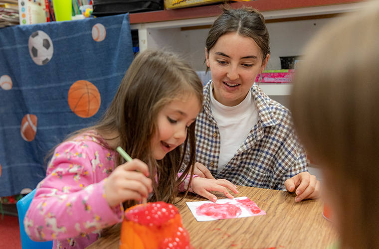 UL Lafayette Art Education student working with a child on an art project blowing colored bubbles through a straw onto paper.