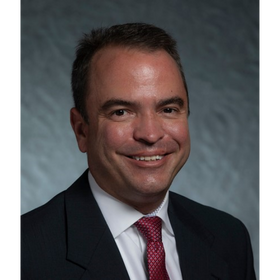 Headshot: Brian West is pictured in a dark-colored blazer, white shirt, and red tie.