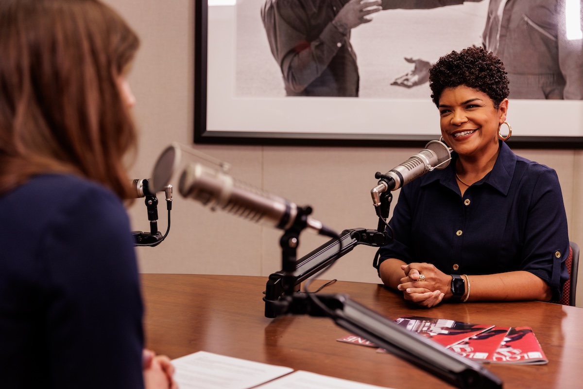 Woman sits at a desk behind a microphone wearing a navy dress