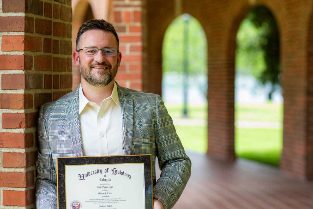 Online MS in Accounting graduate smiles in a white shirt and gray blazer, holding his UL Lafayette master's degree plaque.