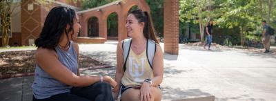 Two University of Louisiana at Lafayette students sit and talk on a bench on campus