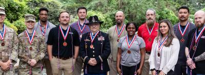 12 people stand on cobblestone pavement in front of green trees and a small pond, each with a red white and blue medallion and cord