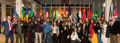 A group of UL Lafayette international students holding up "UL" hand signs in front of a row of flags from around the world