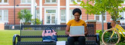 UL Lafayette student sitting on a bench on campus with her laptop in her lap