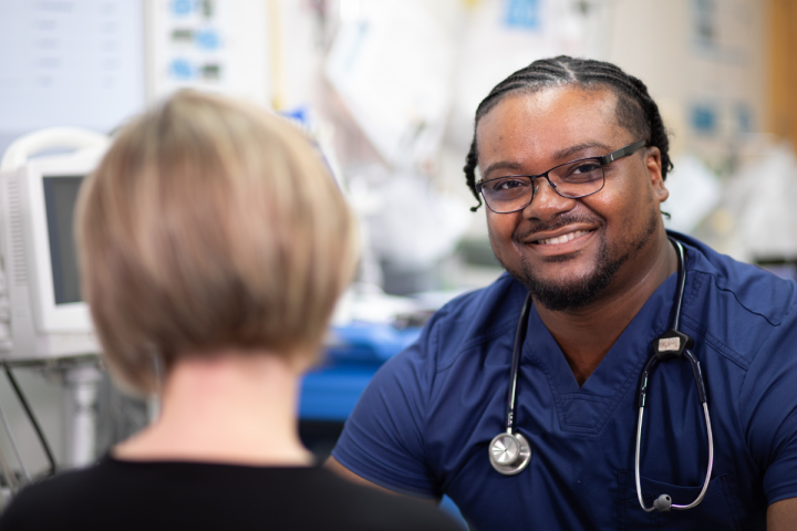 Tristan Lollis is pictured in a hospital exam room wearing blue scrubs. He's smiling toward the camera over the shoulder of a mock patient.