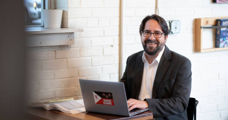 Online student in a blazer sits in coffee shop, working on a laptop that has a Ragin' Cajun/Acadiana Flag sticker.