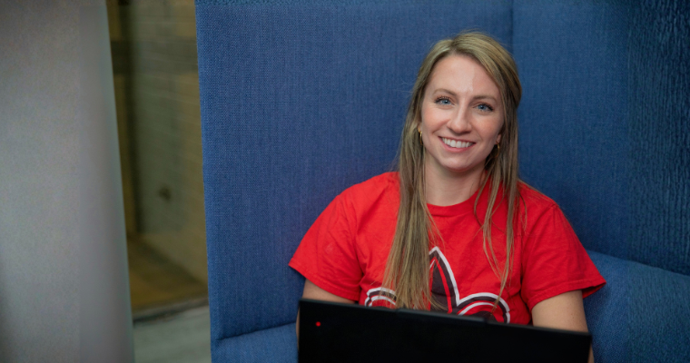 Online student smiles while working on a laptop, wearing a read UL Lafayette t-shirt.