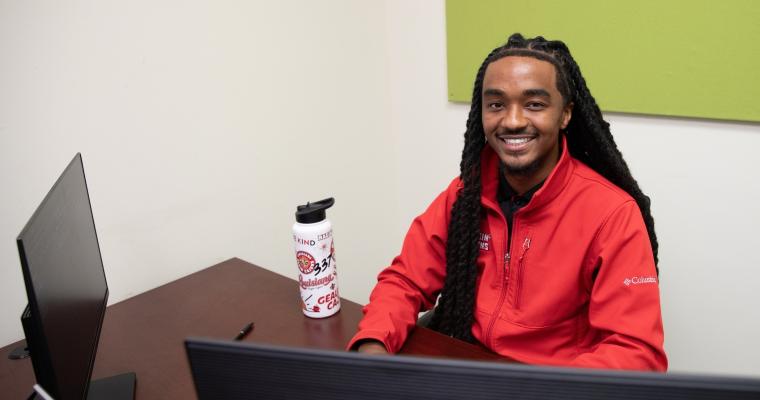 Graduate of UL Lafayette's health services admin degree program smiles while working on a computer. He is wearing a red Ragin' Cajuns quarter-zip.