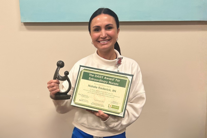RN to BSN online student smiles with her DAISY Award, wearing a white sweater and blue scrub pants.
